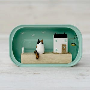 May include: A miniature diorama inside a teal container. It features a white house with a black roof, a calico cat figurine, and a wooden dock. The background has painted seagulls and a fish. The scene is set against a light wood-grain surface.