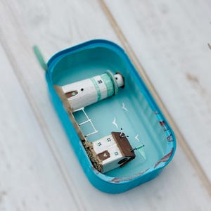 May include: A miniature lighthouse and house scene inside a blue tin can. The lighthouse is white with a green roof and the house is white with a brown roof. The scene is painted with a blue sky and white seagulls. The tin can has a blue lid and a blue bottom.