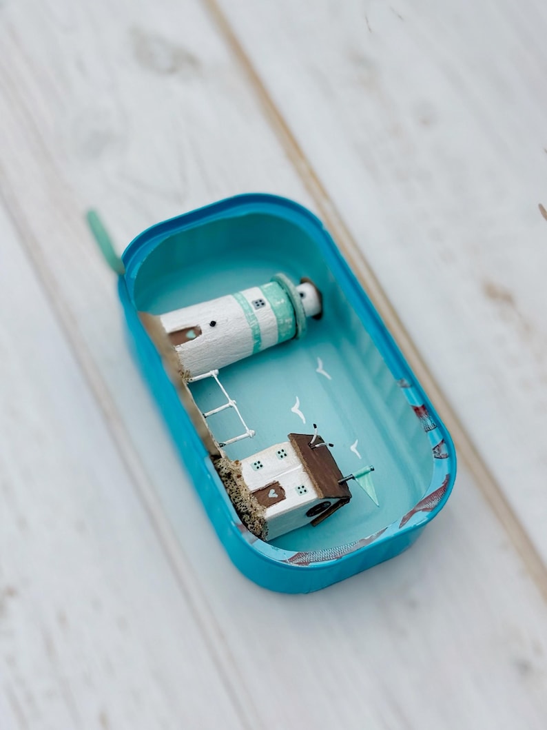 May include: A miniature lighthouse and house scene inside a blue tin can. The lighthouse is white with a green roof and the house is white with a brown roof. The scene is painted with a blue sky and white seagulls. The tin can has a blue lid and a blue bottom.