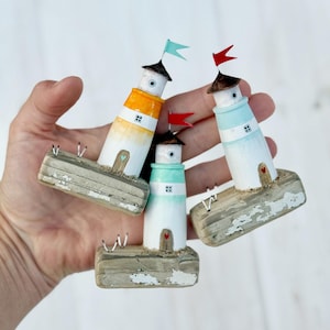 Pu&ograve; includere: Tre figurine di fari in miniatura con corpi dipinti di bianco, arancione e blu, ognuno sormontato da una piccola bandiera. I fari poggiano su basi di legno invecchiato, creando un affascinante tema nautico.