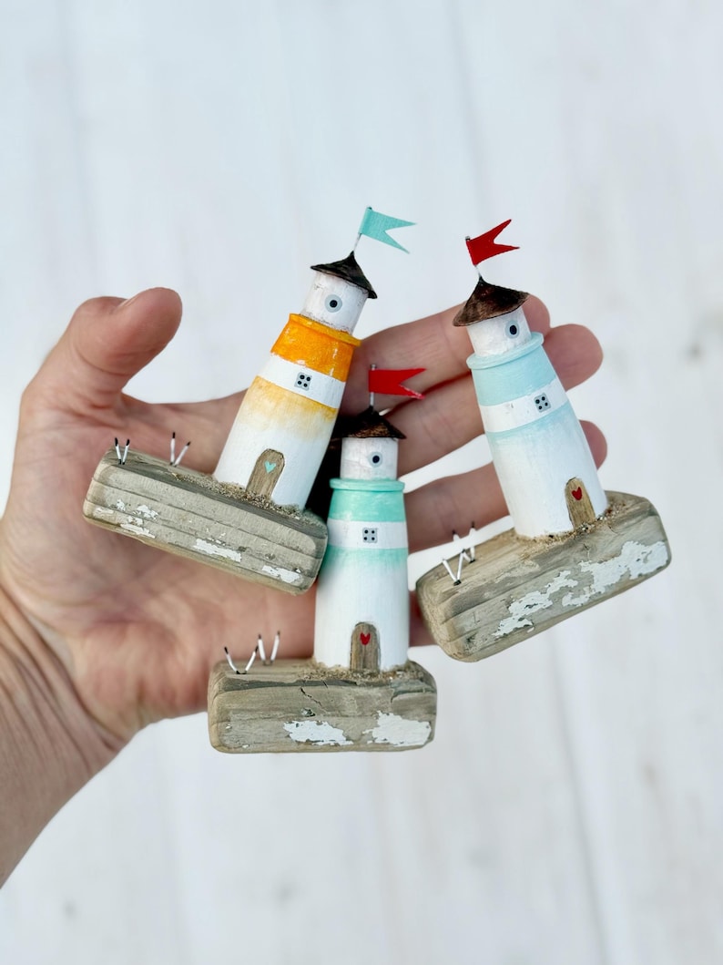 Pu&ograve; includere: Tre figurine di fari in miniatura con corpi dipinti di bianco, arancione e blu, ognuno sormontato da una piccola bandiera. I fari poggiano su basi di legno invecchiato, creando un affascinante tema nautico.