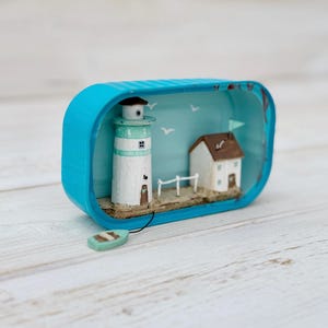 May include: A miniature lighthouse and house scene inside a blue tin can. The lighthouse is white with a green roof and a brown door. The house is white with a brown roof and a blue flag. The scene is set on a sandy beach with a small wooden fence and a blue boat.