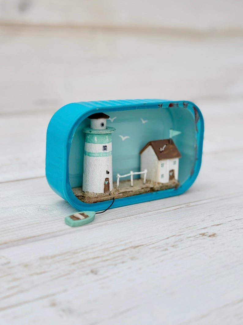 May include: A miniature lighthouse and house scene inside a blue tin can. The lighthouse is white with a green roof and a brown door. The house is white with a brown roof and a blue flag. The scene is set on a sandy beach with a small wooden fence and a blue boat.