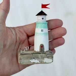 Pu&ograve; includere: Un faro in miniatura in legno, dipinto di bianco e verde acqua, con una bandiera rossa. Il faro presenta una piccola porta con un ritaglio a forma di cuore e poggia su una base in legno invecchiato. Due piccole figure di uccelli sono sulla base.