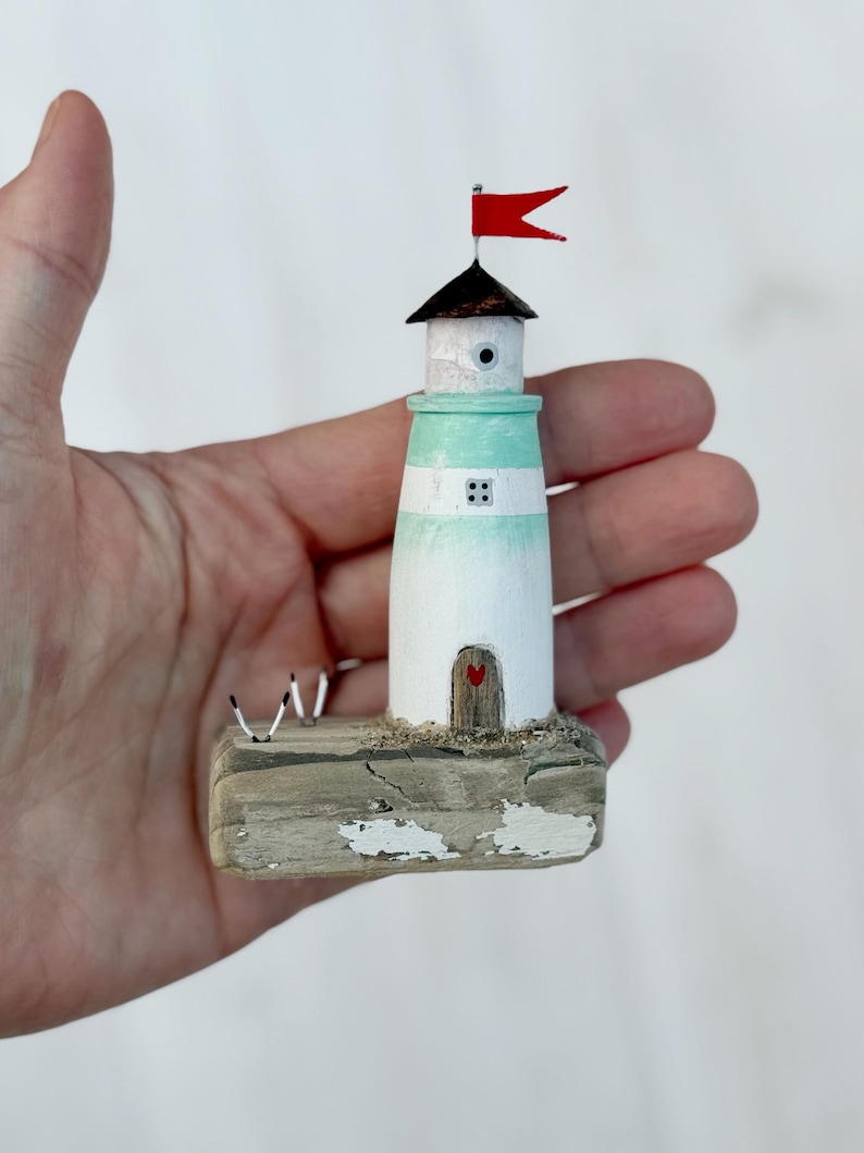 Pu&ograve; includere: Un faro in miniatura in legno, dipinto di bianco e verde acqua, con una bandiera rossa. Il faro presenta una piccola porta con un ritaglio a forma di cuore e poggia su una base in legno invecchiato. Due piccole figure di uccelli sono sulla base.