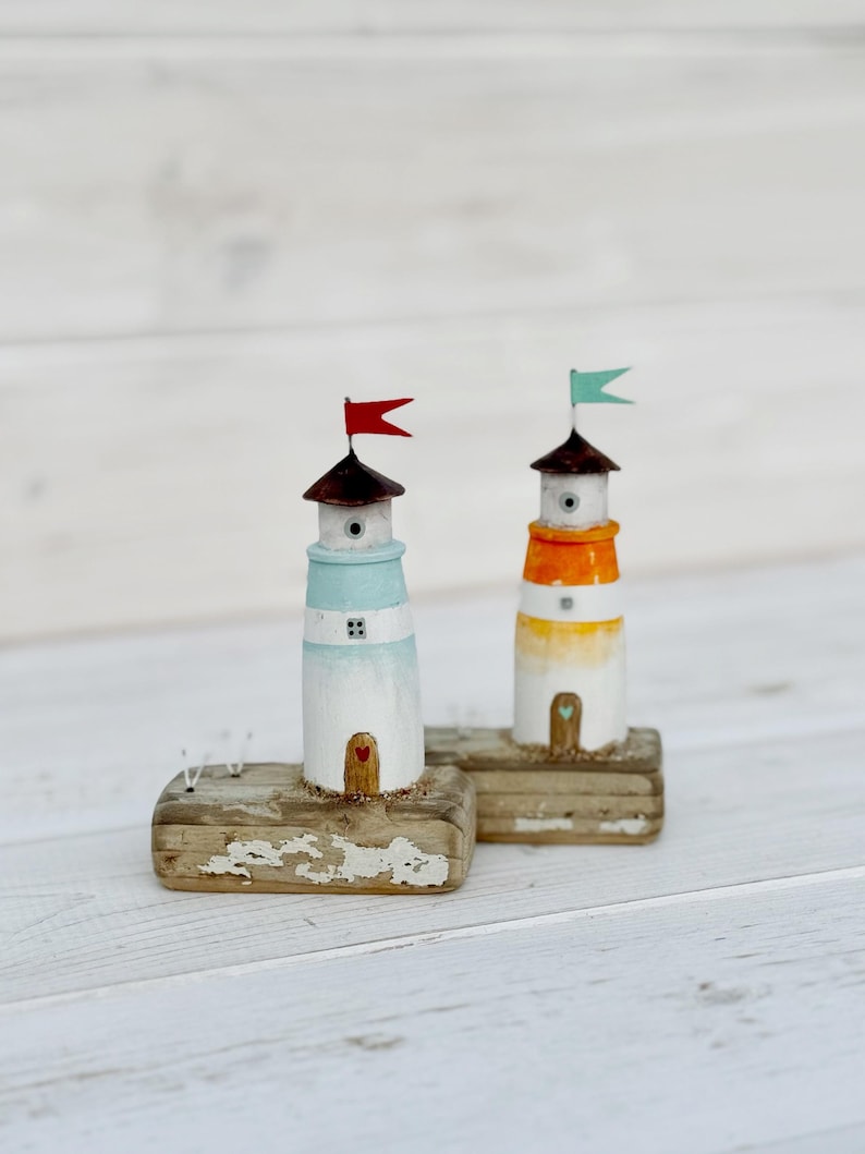 Pu&ograve; includere: Due figurine di fari in legno in miniatura. Uno &egrave; dipinto di bianco e azzurro con una bandiera rossa, l'altro &egrave; bianco e arancione con una bandiera verde. Entrambi hanno tetti marroni e poggiano su basi di legno invecchiato.
