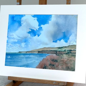 May include: Watercolour painting of a coastal landscape featuring a blue sea, brown cliffs, and a cloudy sky. The artwork is presented in a white mount and frame, displayed on a wooden easel.