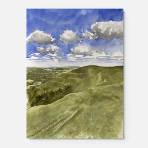 May include: Watercolour painting depicting a landscape with undulating green hills beneath a blue sky dotted with white clouds. The artwork portrays a tranquil outdoor scene, with a distant view of a town or village.