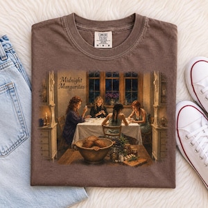 May include: A brown t-shirt featuring a graphic print of four individuals sitting at a table in a dimly lit room. The text Midnight Margaritas is printed in the upper left corner of the graphic. The shirt has a crew neckline and a Comfort Colors label.