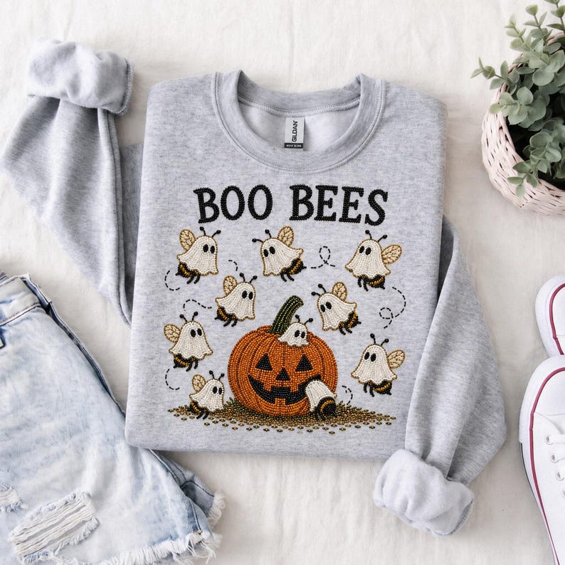 K&ouml;nnte beinhalten: Ein hellgraues Sweatshirt mit Rundhalsausschnitt und einer Stickerei, die einen gro&szlig;en orangefarbenen K&uuml;rbis zeigt, umgeben von kleinen Bienen in wei&szlig;en Geisterkost&uuml;men. Der Text "BOO BEES" ist in Schwarz &uuml;ber dem Motiv gestickt.