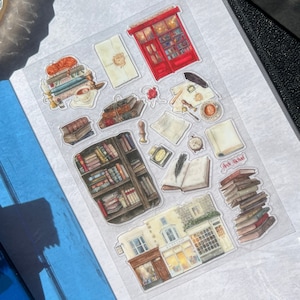 May include: A sheet of clear stickers with illustrations of books, a bookshelf, a storefront, and other book-related items. The stickers are printed with a vintage aesthetic and feature a red, white, and blue color scheme.