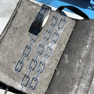 May include: A black and silver washi tape roll with a chain design sits on a gray textured surface, next to several silver chain stickers. The tape and stickers are displayed on an open, dark gray journal with a black strap.