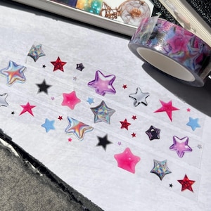 Puede incluir: Una hoja de cinta adhesiva transparente con pegatinas de estrellas holográficas en rosa, morado, azul y plateado. Las estrellas están dispuestas en filas en la cinta.