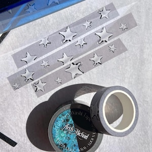 Puede incluir: Cinta washi gris con un diseño de estrellas, junto con tiras de la misma cinta. El rollo de cinta tiene el texto "Arch Michael" y "shoparchmichael.com". La cinta está sobre una superficie blanca.