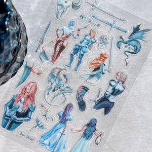 May include: A clear sticker sheet featuring fantasy-themed illustrations. The designs include figures in armor, swords, dragons, and ethereal creatures in shades of blue, white, and brown. The text "Arch Michael" is visible on the sheet.