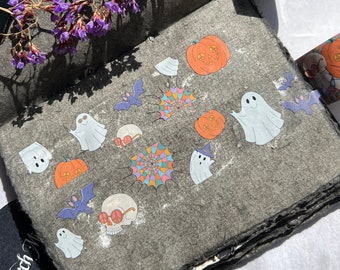 May include: A black scrapbook with a variety of Halloween themed stickers, including ghosts, pumpkins, bats, and a skull. The stickers are arranged in a whimsical pattern on the dark gray paper.