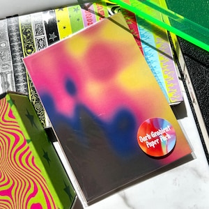 May include: A stack of paper packs with a dark gradient design in pink, yellow, and blue. The packs are labeled "Dark Gradient Paper Pack" and are surrounded by other stationery items with colorful patterns and neon green accents.