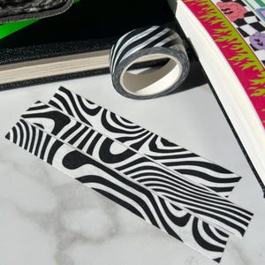 Puede incluir: Un rollo de cinta adhesiva washi a rayas blancas y negras se encuentra junto a dos tiras con el mismo patrón. Un cuaderno negro y un diario colorido también están en la imagen. La cinta y las tiras tienen un diseño ondulado, similar al de una cebra.