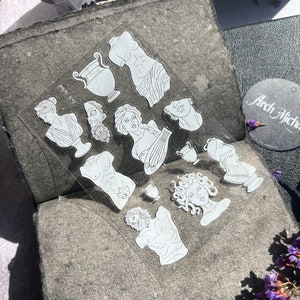 May include: A clear sticker sheet featuring white line art illustrations of classical Greek busts, torsos, and vases. The sticker sheet is displayed on a textured gray surface, with the text "Arch Michael" visible on a tag.
