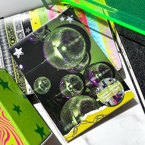 Puede incluir: Una colección de artículos de papelería en verde neón y negro. Los artículos incluyen una caja con un diseño de remolino rosa y verde, una bandeja verde transparente y un paquete de papel con gráficos de bolas de discoteca. El paquete de papel tiene el texto "Chaos Theory Paper Pack".