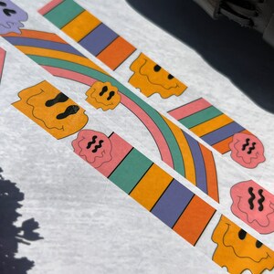 May include: Three colorful washi tape rolls with a rainbow design and a dripping smiley face. The tape is on a white background.