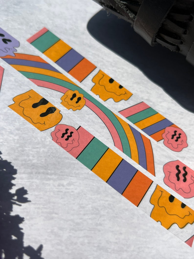 May include: Three colorful washi tape rolls with a rainbow design and a dripping smiley face. The tape is on a white background.