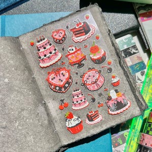 May include: An open grey journal with decorative cake stickers. The stickers feature various pink, red, and black cakes with cherries, strawberries, and other embellishments. The journal is surrounded by colourful washi tape rolls.