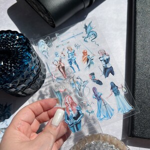 May include: A clear sticker sheet featuring fantasy-themed illustrations. The stickers depict figures in armor, dragons, swords, and other medieval fantasy elements. The color palette includes blues, whites, and browns. The sticker sheet is held by a hand.
