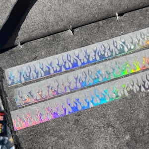Puede incluir: Primer plano de tres tiras de pegatinas holográficas de llamas. Las pegatinas tienen un diseño de llama repetitivo que cambia de color de azul a morado a rojo. Se colocan sobre un fondo gris texturizado.
