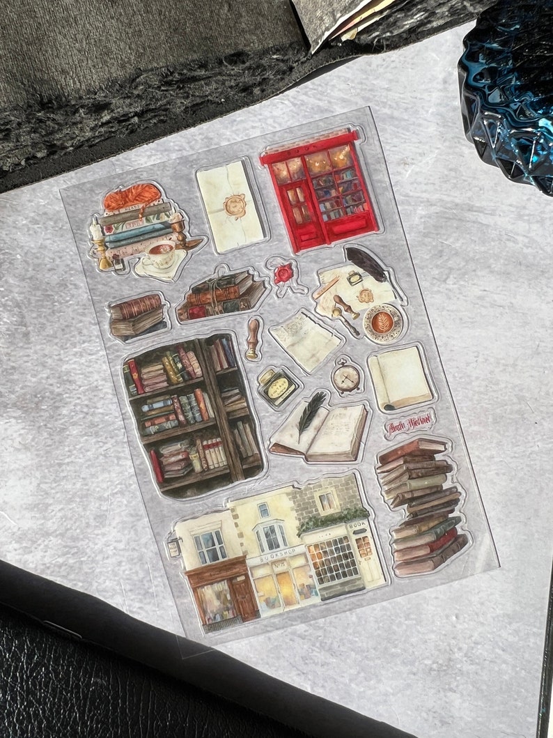 May include: A sheet of clear stickers with illustrations of a bookstore, books, and other book-related items. The stickers include a red storefront with the words "Book Nook" above the door, a stack of books with the words "Read More!" on top, and a bookshelf filled with books. The stickers are perfect for adding a touch of whimsy to journals, planners, and other paper crafts.