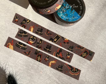 May include: Three rolls of washi tape with a black and gold glitter background. The tape features a repeating pattern of a hand holding a crystal ball, a snake, a candle, and a tarot card. The tape is labelled "Shop Vranai" and "Washi Tape 15mm 5m".