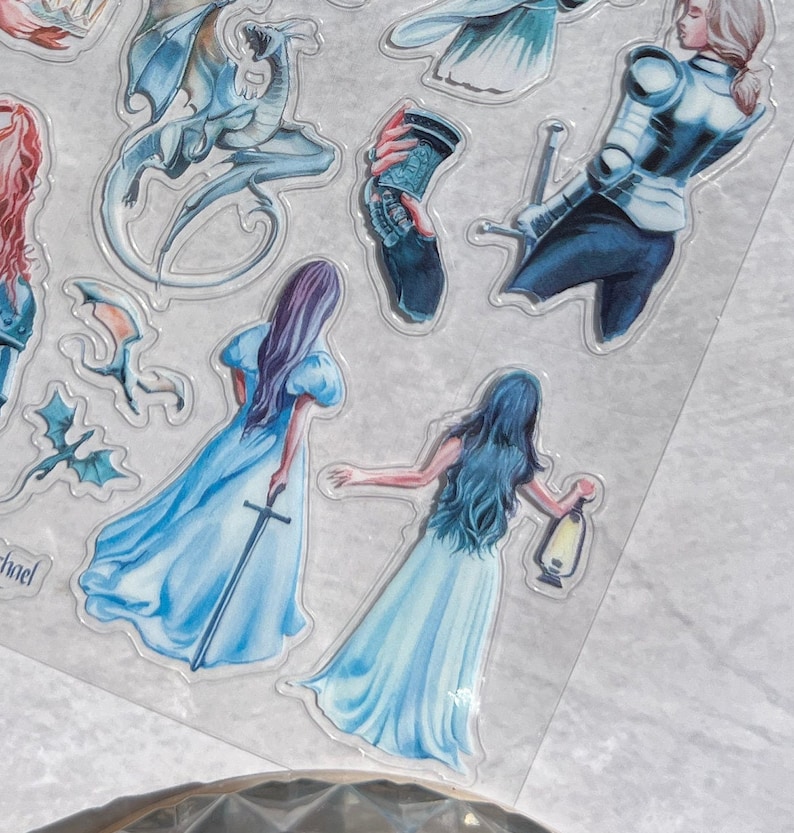 May include: A sheet of fantasy-themed stickers featuring illustrations of dragons, women in flowing gowns, a knight, and other mystical elements. The stickers are in shades of blue, purple, and white, with a transparent border.