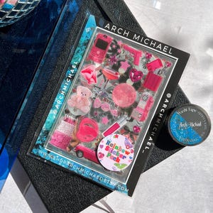 May include: A sheet of pink and red stickers with a variety of designs, including a teddy bear, a lipstick, a phone, a heart, a star, a gun, a cake, a car, and a martini glass. The stickers are packaged in a clear plastic sheet with the text "Rainbow Sticker Pack" and "@ARCHMICHAEL_" printed on it. The sheet is sitting on a black surface with a roll of washi tape.