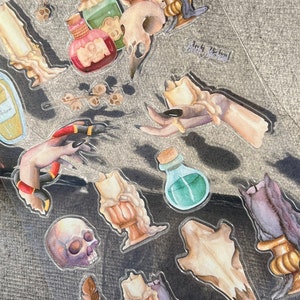 May include: A sheet of clear stickers with a witchy theme. The stickers feature a hand holding a candle, a skull, a potion bottle, and a snake. The stickers are perfect for adding a touch of magic to your planner, journal, or scrapbook.