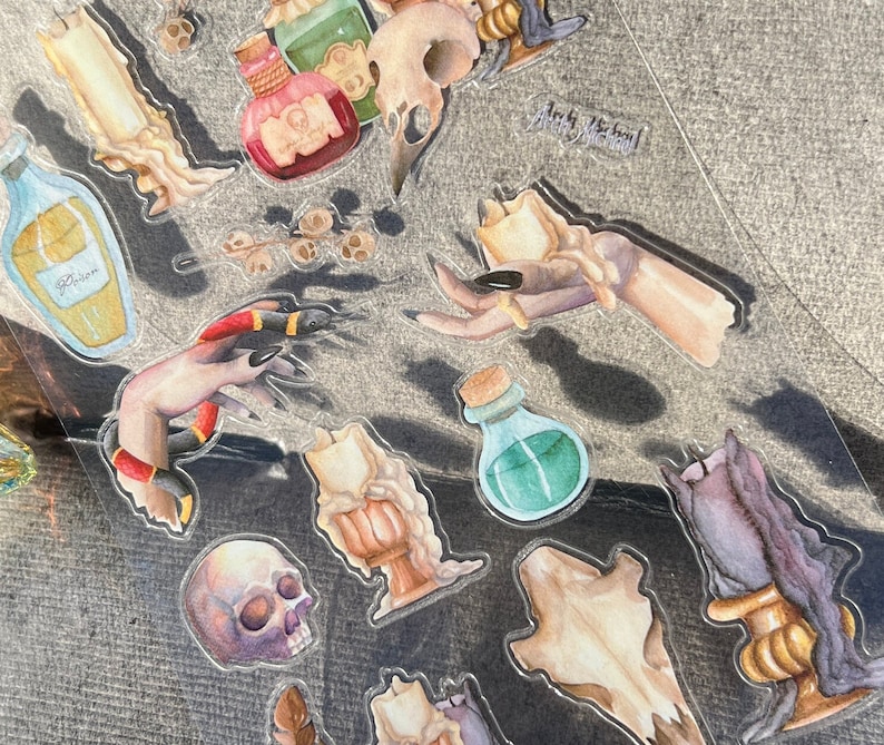 May include: A sheet of clear stickers with a witchy theme. The stickers feature a hand holding a candle, a skull, a potion bottle, and a snake. The stickers are perfect for adding a touch of magic to your planner, journal, or scrapbook.