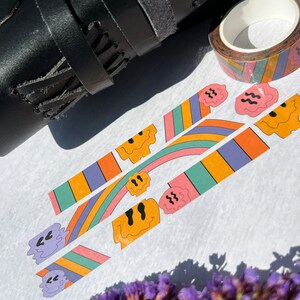May include: Three rolls of colorful washi tape with a rainbow and smiley face design. The tape is on a white surface with a black leather strap and purple flowers.
