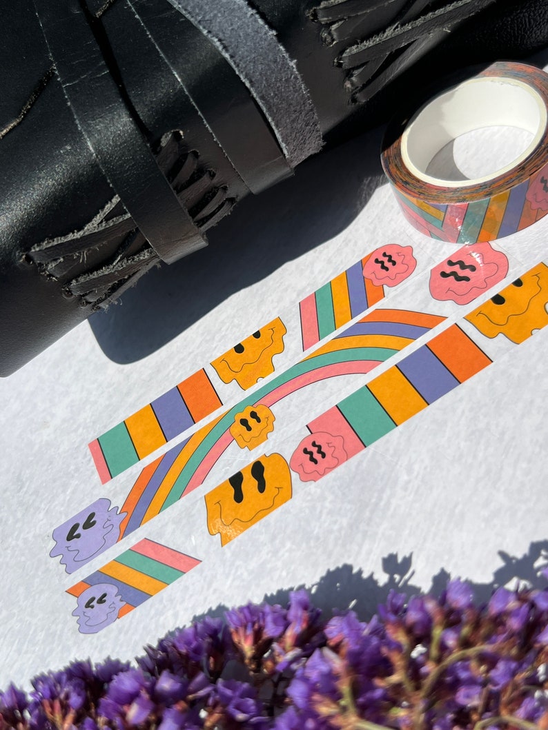 May include: Three rolls of colorful washi tape with a rainbow and smiley face design. The tape is on a white surface with a black leather strap and purple flowers.