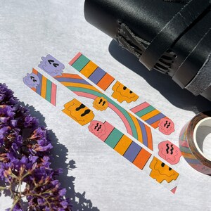May include: Three rolls of colorful washi tape with a repeating pattern of smiling faces and rainbow stripes. The tape is on a white surface with a black leather notebook and a roll of brown washi tape in the background.