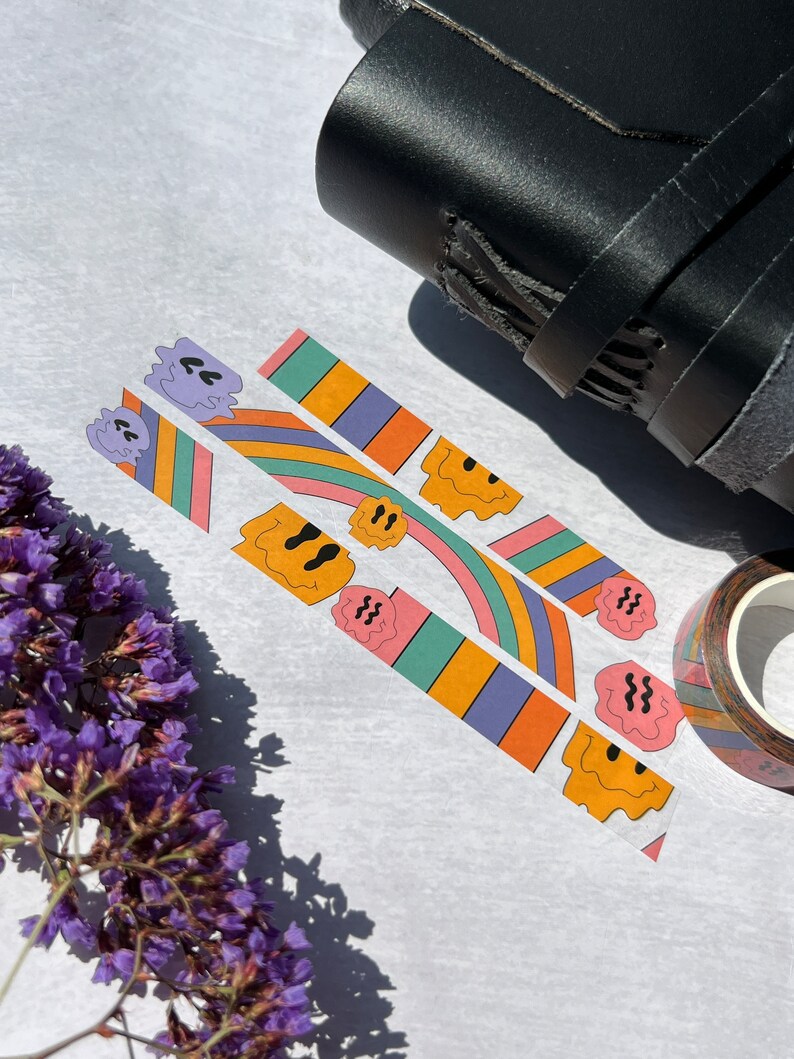 May include: Three rolls of colorful washi tape with a repeating pattern of smiling faces and rainbow stripes. The tape is on a white surface with a black leather notebook and a roll of brown washi tape in the background.