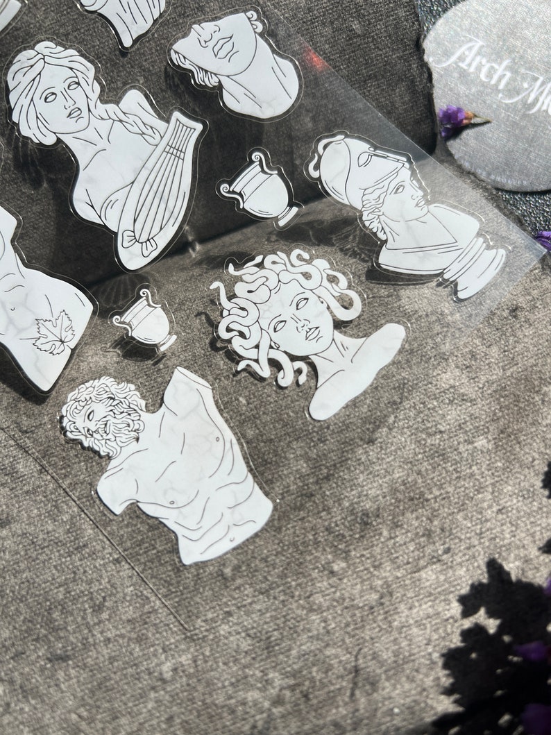 May include: A collection of white, die-cut stickers featuring classical Greek and Roman sculptures. The stickers include busts, figures, and musical instruments. The stickers are on a clear backing, with the text "Arch Me" visible in the upper right corner.