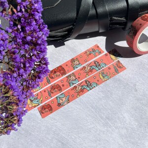 K&ouml;nnte beinhalten: Drei Rollen rosa Washi-Tape mit einem sich wiederholenden Muster einer Frau, die auf einem zunehmenden Mond mit Sternen reitet, und einem Farbschema in Rot, Orange und Gelb.