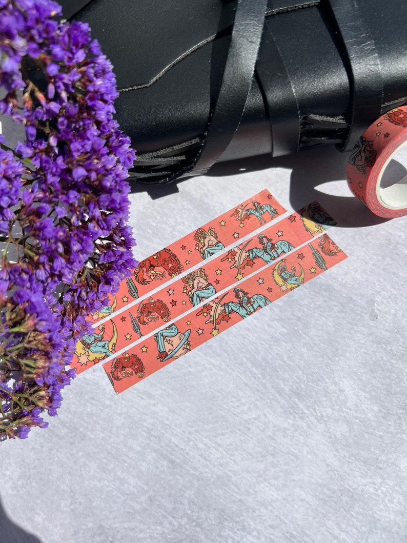 K&ouml;nnte beinhalten: Drei Rollen rosa Washi-Tape mit einem sich wiederholenden Muster einer Frau, die auf einem zunehmenden Mond mit Sternen reitet, und einem Farbschema in Rot, Orange und Gelb.
