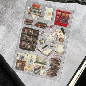 May include: A sheet of clear stickers featuring illustrations of books, a bookstore, a red mailbox, and other bookish elements. The stickers are perfect for adding a touch of whimsy to journals, planners, and other paper crafts.