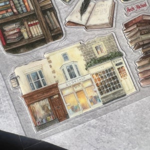 May include: A watercolor illustration of a bookstore with two storefronts. The storefront on the left says "The Book Shop" and the storefront on the right says "Bookshop No. 75".