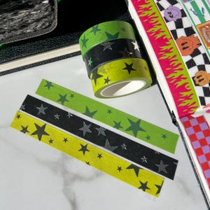 Puede incluir: Cintas washi decorativas en verde, negro y amarillo, adornadas con patrones de estrellas plateadas. Las cintas están apiladas y acompañadas de tiras a juego, ideales para manualidades y journaling.
