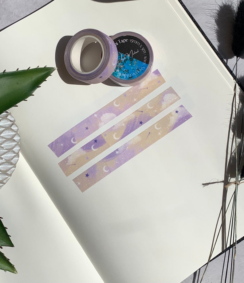 May include: Three rolls of washi tape with a purple and cream color scheme. The tape features a repeating pattern of clouds, stars, and crescent moons. The tape is labeled "Washi Tape" and has a small logo.
