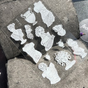 May include: A clear sheet of stickers featuring white line art of classical sculptures, busts, and vases. The stickers include images of a torso, a woman with a lyre, and Medusa. The text "Arch-Yktied" is printed on the sheet.