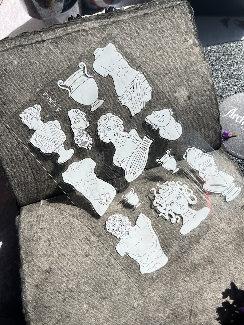 May include: A clear sheet of stickers featuring white line art of classical sculptures, busts, and vases. The stickers include images of a torso, a woman with a lyre, and Medusa. The text "Arch-Yktied" is printed on the sheet.