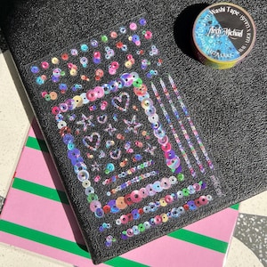 May include: A clear sticker sheet featuring iridescent, multi-colored sequin-style circular, heart, and star shapes, alongside a roll of Arch Michael washi tape measuring 15mm by 10m, positioned on a black textured surface over a pink and green striped base.