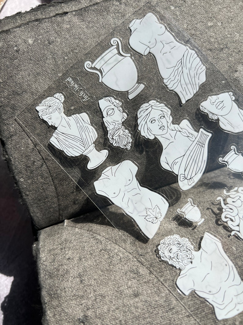 May include: Clear sticker sheet featuring white line art of classical Greek statues, busts, and vases. The text "Arch Method" is printed on the sticker sheet. The stickers are on a gray surface.
